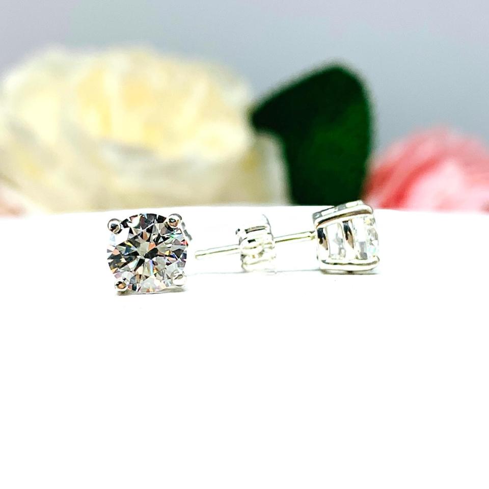 A pair of silver stud earrings set with round brilliant cut 'near colorless or 'G color' cubic zirconias.  The four prong basket earrings are on a white surface with the left earring facing forward and the right earring facing to the right to showcase the brilliant cutting of the czs, center post and butterfly friction clasp.