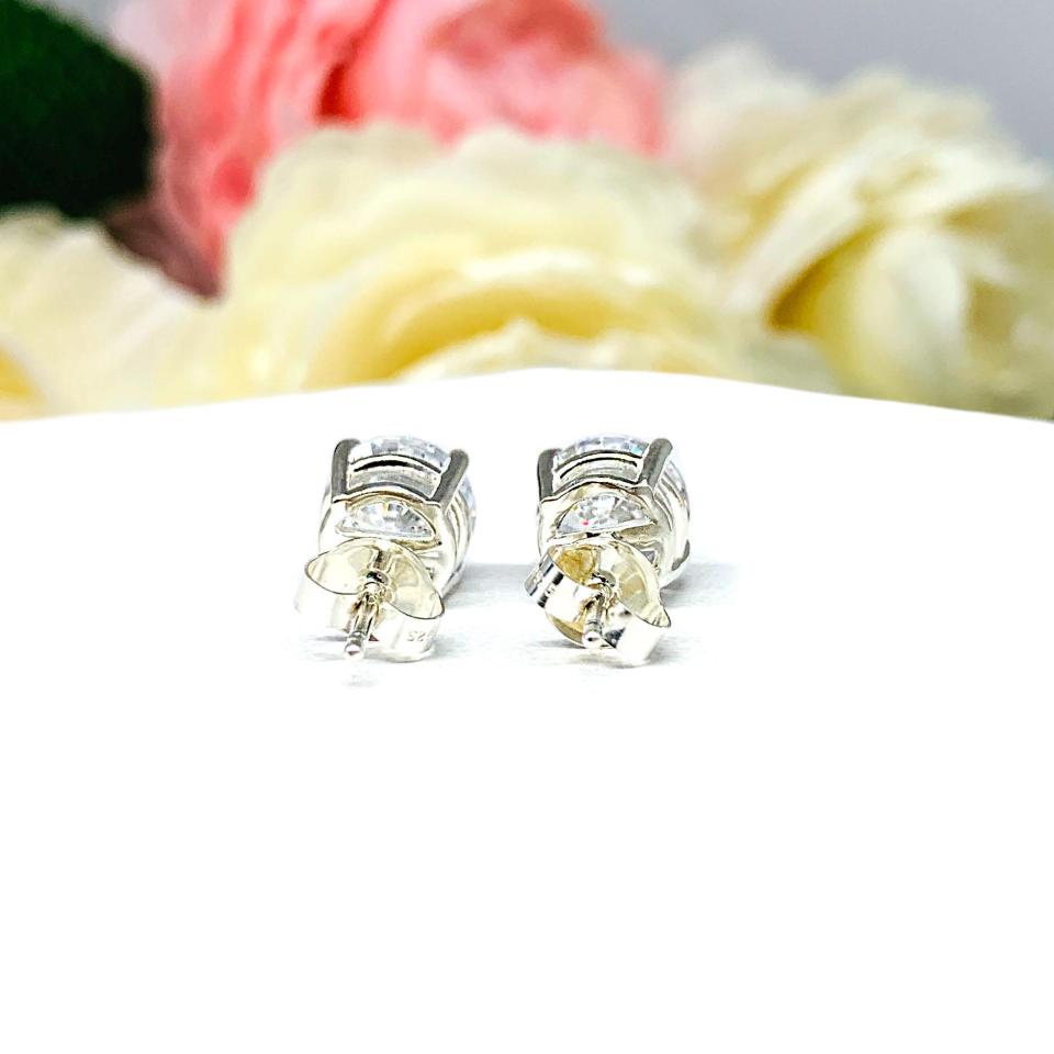 A pair of silver stud earrings set with round brilliant cut cubic zirconias.  The four prong basket earrings are on a white surface with both earrings facing toward the back to showcase the center post and butterfly friction clasps.