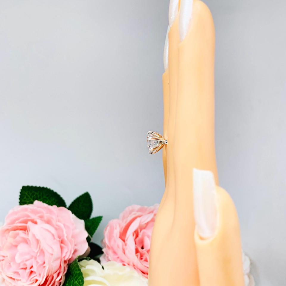Mannequin hand with long white shimmery nails, displayed against a soft white-gray background with pink and white floral accents. The hand is wearing a 10mm near colorless (G/H color) round brilliant cut cubic zirconia solitaire engagement ring in a classic 6-prong setting. The setting is simple yet elegant, allowing the center stone to take center stage.