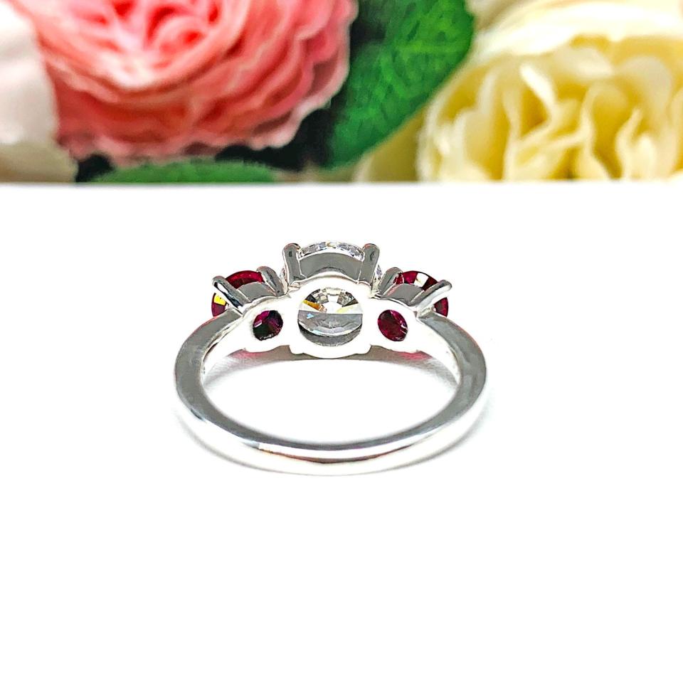 Sterling silver 3-stone ring with a round brilliant cut colorless (D color) cubic zirconia center flanked by two deep red lab-created rubies. The ring is set on a white surface with a background of pink and white flowers.
