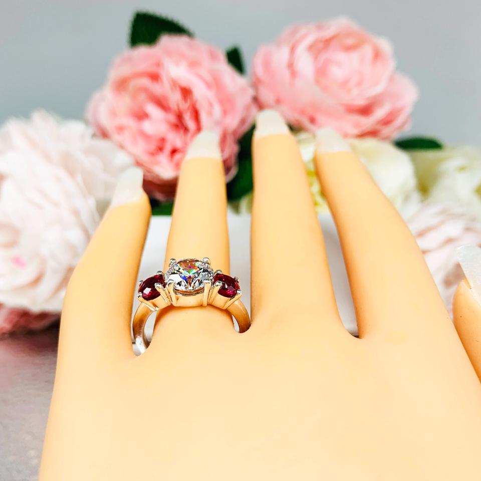 Mannequin hand with long white shimmery nails, displayed against a soft white-gray background with pink and white floral accents. The hand is wearing a sterling silver 3-stone ring with a 9mm near colorless round brilliant cut cubic zirconia center that radiates icy sparkle, flanked by two 5mm round deep red lab-created rubies.