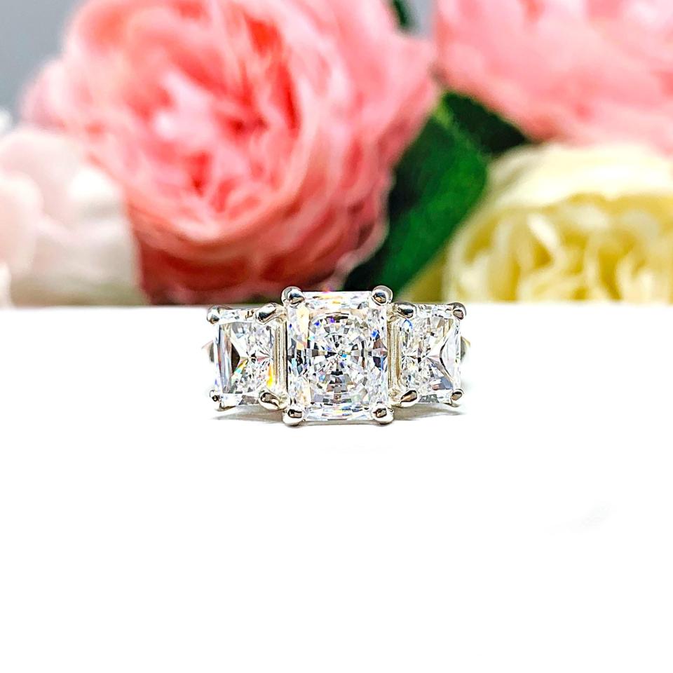Sterling silver 3-stone engagement ring featuring a 9x7mm radiant cut cubic zirconia center stone with two 7x5mm radiant CZ side stones. The highly polished wide band tapers for comfort. Displayed on a white surface with soft floral background.