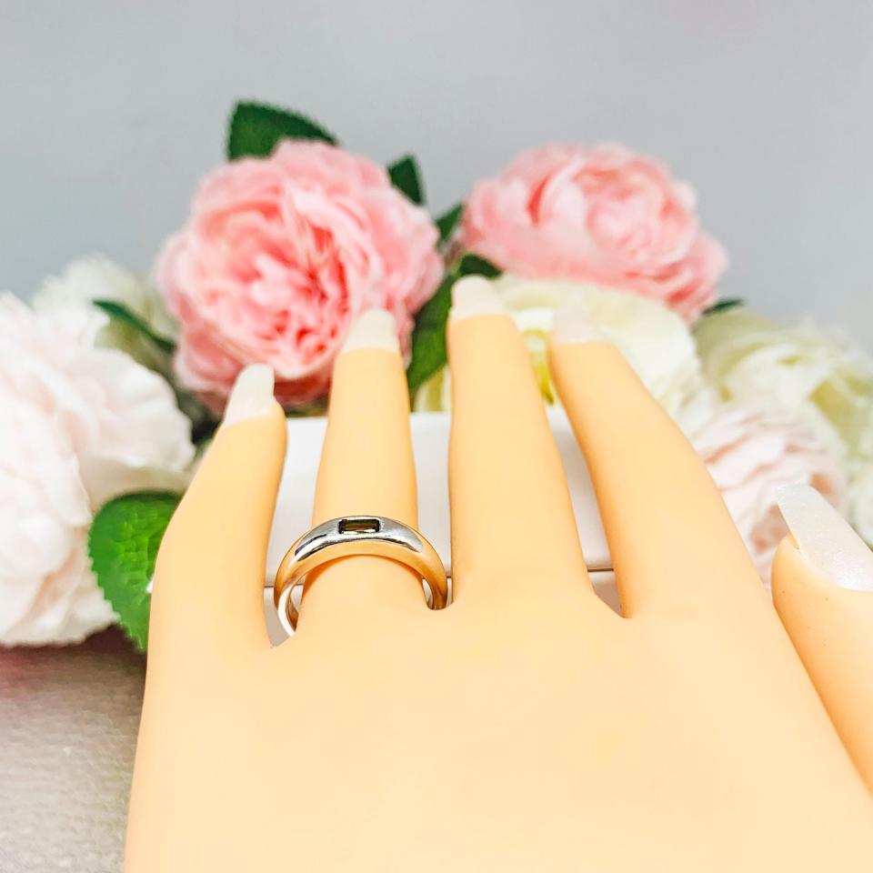Mannequin hand with long white shimmery nails, displayed against a soft white-gray background with pink and white floral accents. The hand is wearing a sterling silver band featuring a 5×3 mm emerald‑cut green tourmaline set flush in a polished band.