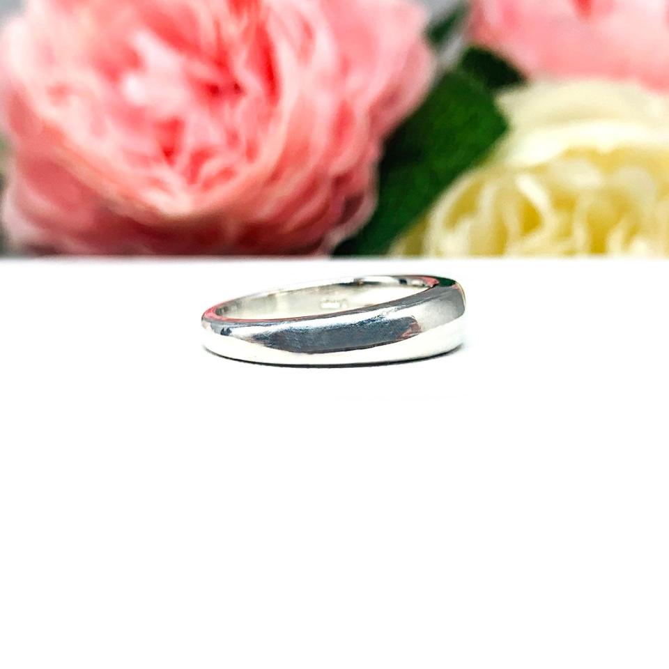 Sterling silver band featuring a 5×3 mm emerald‑cut green tourmaline set flush in a polished band, displayed on a white surface with soft pink and cream flowers blurred in the background.