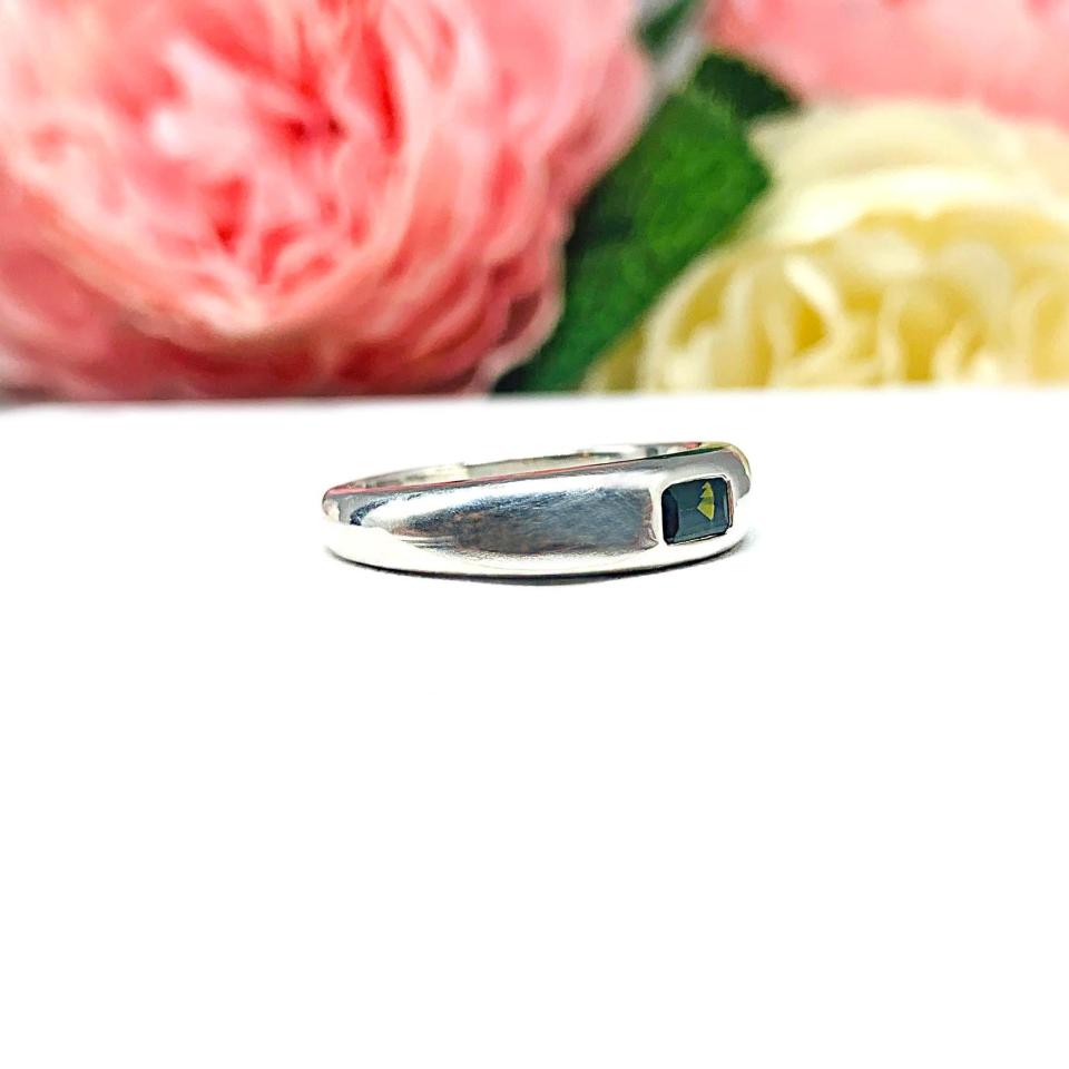 Sterling silver band featuring a 5×3 mm emerald‑cut green tourmaline set flush in a polished band, displayed on a white surface with soft pink and cream flowers blurred in the background.