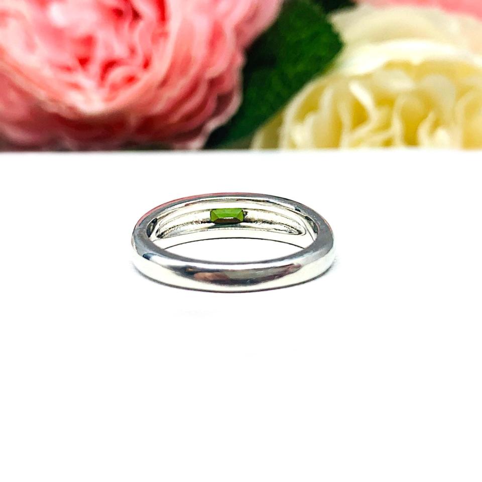 Sterling silver band featuring a 5×3 mm emerald‑cut green tourmaline set flush in a polished band, displayed on a white surface with soft pink and cream flowers blurred in the background.