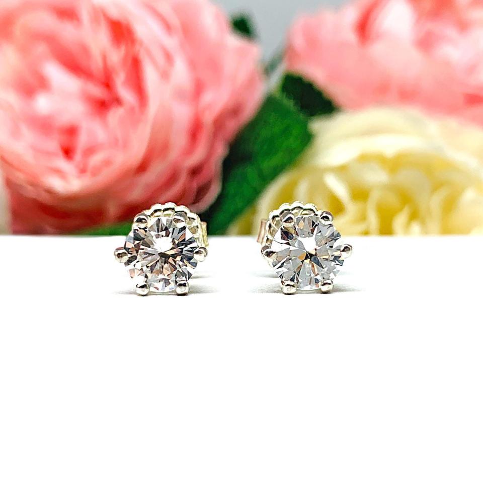 A pair of sterling silver six prong stud earrings set with round brilliant lab white sapphires. The earrings have center posts and butterfly clasps and are displayed on a white surface with a floral background.