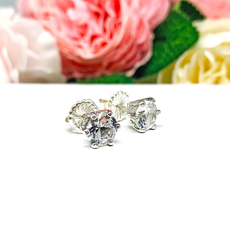 A pair of sterling silver six prong stud earrings set with round brilliant lab white sapphires. The earrings have center posts and butterfly clasps and are displayed on a white surface with a floral background.
