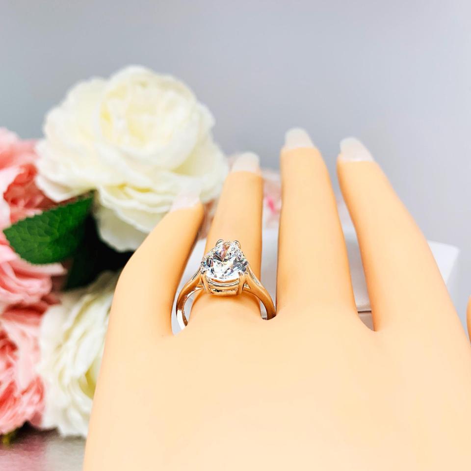 Mannequin hand with long white shimmery nails, displayed against a soft white-gray background with pink and white floral accents. The hand is wearing an elegant 4-prong sterling silver cathedral engagement ring set featuring a 13x9mm near colorless precision-cut pear cubic zirconia. Displayed on a white surface with a floral background.