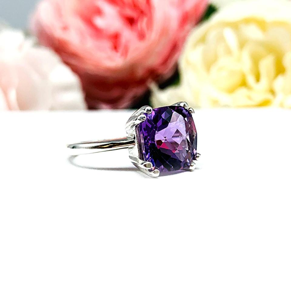 Sterling silver solitaire ring featuring a 10mm checkerboard cushion-cut vibrant African amethyst gemstone. The vibrant purple gem ring is set against a soft floral background that enhances the ring’s natural elegance.