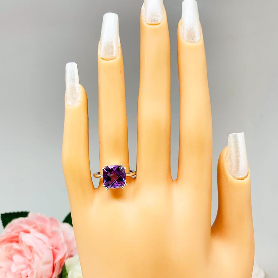 Mannequin hand with long white shimmery nails, displayed against a soft white-gray background with pink and white floral accents. The hand is wearing a sterling silver solitaire ring featuring a 10mm checkerboard cushion-cut vibrant African amethyst gemstone. The vibrant purple gem ring is set against a soft floral background that enhances the ring’s natural elegance.