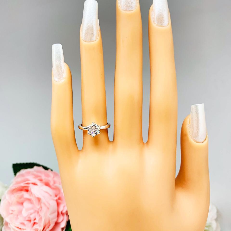 Mannequin hand with long white shimmery nails, displayed against a soft white-gray background with pink and white floral accents. The hand is wearing a sterling silver round-cut DEF moissanite 6-prong engagement ring. Displayed on a white surface with pink and white flowers in the background.