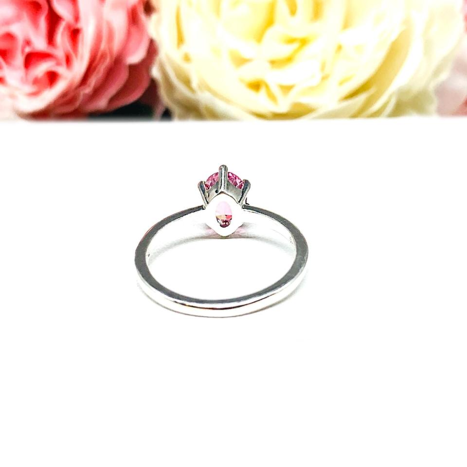 Sterling silver ring with an 8x6mm oval lab created padparadscha sapphire set in a 6-prong setting set against a white surface with soft-focus pink and white flowers in the background.