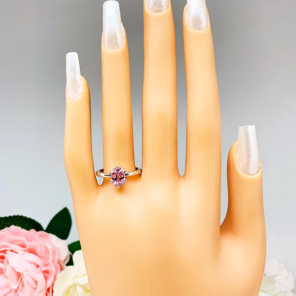 Mannequin hand with long white shimmery nails, displayed against a soft white-gray background with pink and white floral accents. The hand is wearing a sterling silver ring with an 8x6mm oval lab created padparadscha sapphire set in a 6-prong setting.