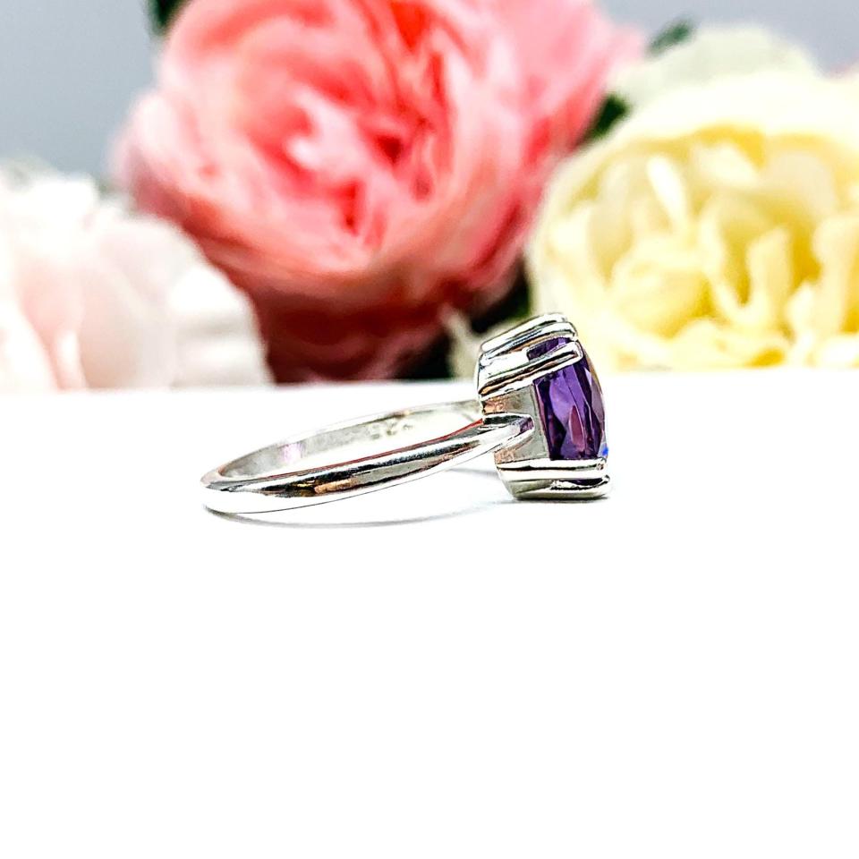 Sterling silver solitaire ring featuring a 10mm checkerboard cushion-cut vibrant African amethyst gemstone. The vibrant purple gem ring is set against a soft floral background that enhances the ring’s natural elegance.