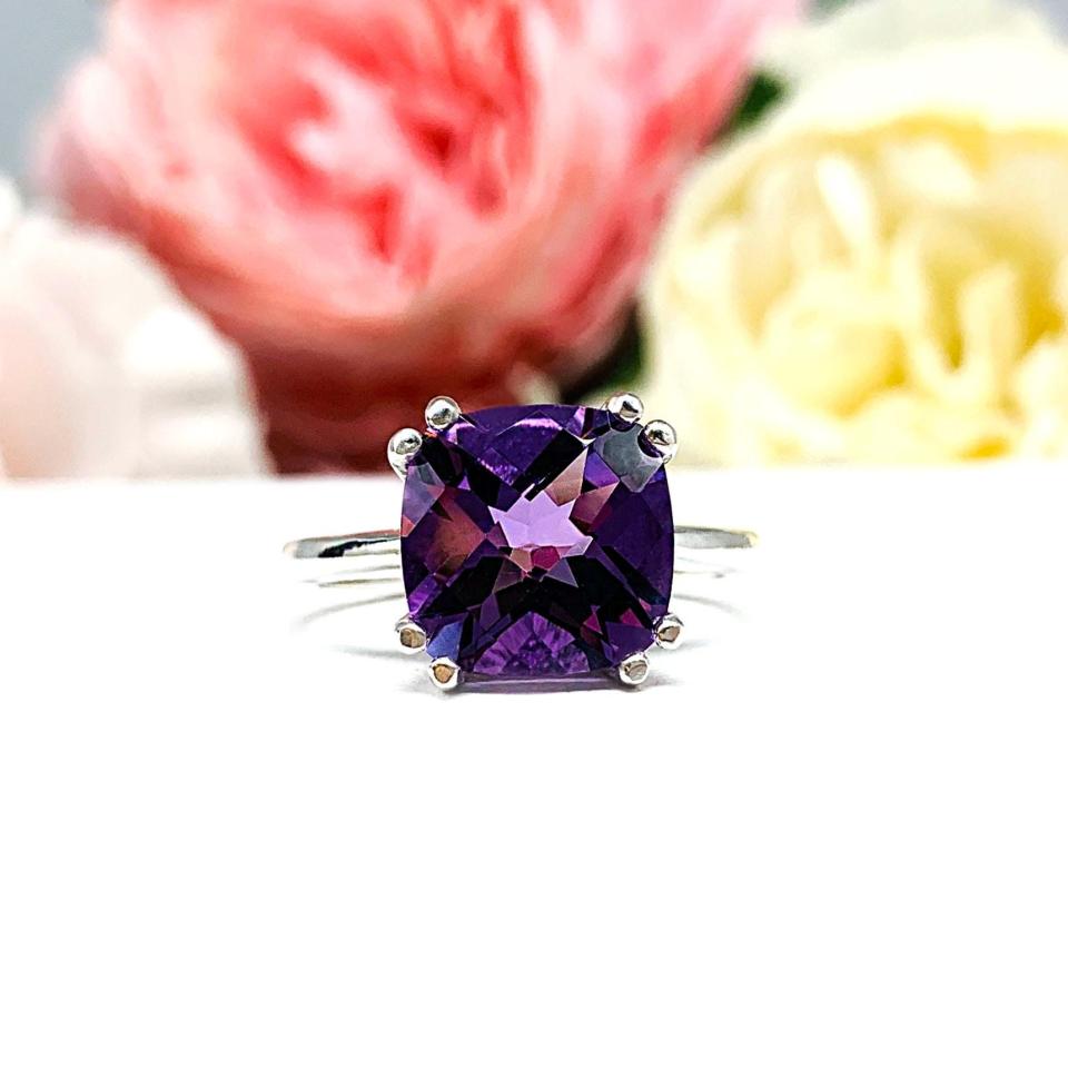 Sterling silver solitaire ring featuring a 10mm checkerboard cushion-cut vibrant African amethyst gemstone. The vibrant purple gem ring is set against a soft floral background that enhances the ring’s natural elegance.