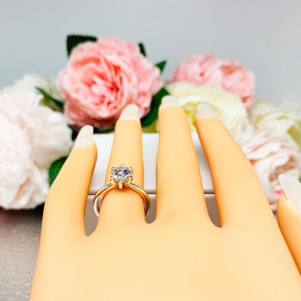 Mannequin hand with long white shimmery nails, displayed against a soft white-gray background with pink and white floral accents. The hand is wearing a sterling silver round-cut DEF moissanite 6-prong engagement ring. Displayed on a white surface with pink and white flowers in the background.