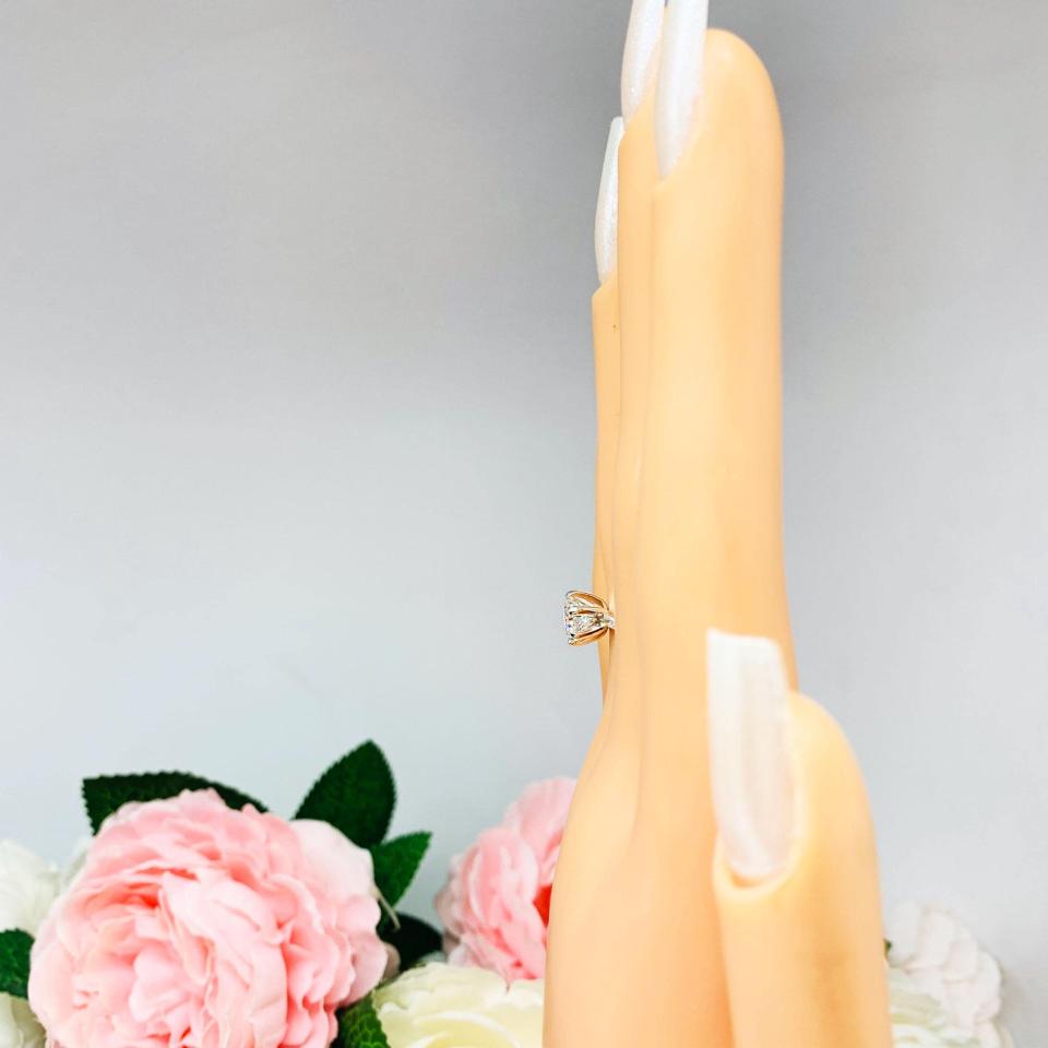 Mannequin hand with long white shimmery nails, displayed against a soft white-gray background with pink and white floral accents. The hand is wearing a sterling silver round-cut DEF moissanite 6-prong engagement ring. Displayed on a white surface with pink and white flowers in the background.