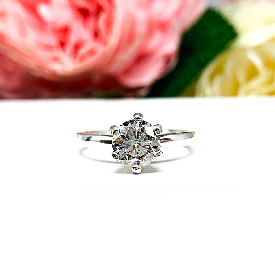 Sterling silver round-cut DEF moissanite 6-prong engagement ring. Displayed on a white surface with pink and white flowers in the background.