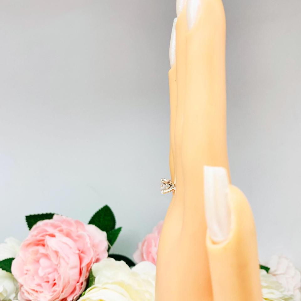Mannequin hand with long white shimmery nails, displayed against a soft white-gray background with pink and white floral accents. The hand is wearing a solitaire engagement ring with a round brilliant DEF moissanite. The setting is a classic four-prong design, and the band is a simple, sterling silver band. The ring is displayed on a white background with blurred pink and yellow flowers in the background.