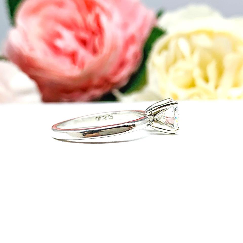 A solitaire engagement ring with a round brilliant DEF moissanite. The setting is a classic four-prong design, and the band is a simple, sterling silver band. The ring is displayed on a white background with blurred pink and yellow flowers in the background.