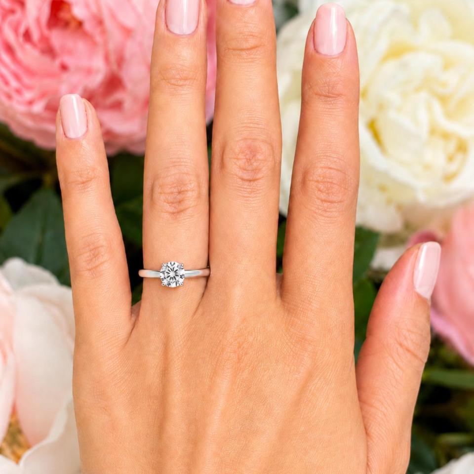 Woman's hand wearing a sterling silver 6.5mm round-cut 4-prong moissanite engagement ring. Model image for size and styling reference — not a photo of the actual ring.