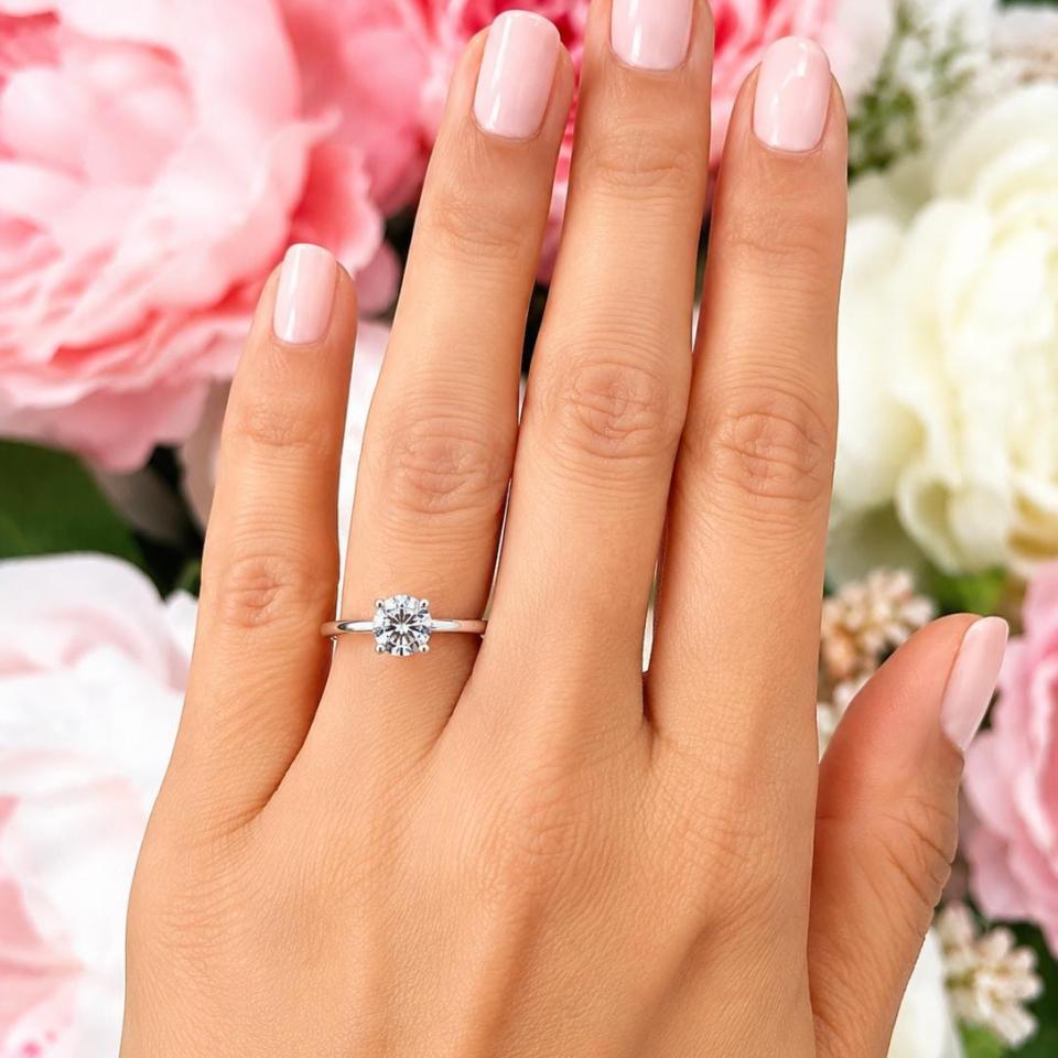 Woman's hand wearing a sterling silver 7mm round-cut 4-prong moissanite engagement ring. Model image for size and styling reference — not a photo of the actual ring.