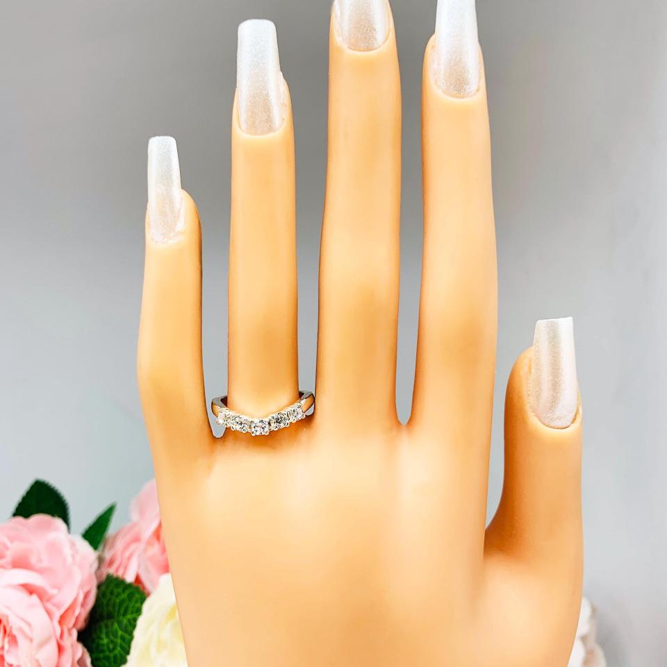 Mannequin hand with long white shimmery nails, displayed against a soft white-gray background with pink and white floral accents. The hand is wearing a sterling silver 5-stone curved wedding band featuring 3mm round precision faceted near colorless cz stones in prong settings.