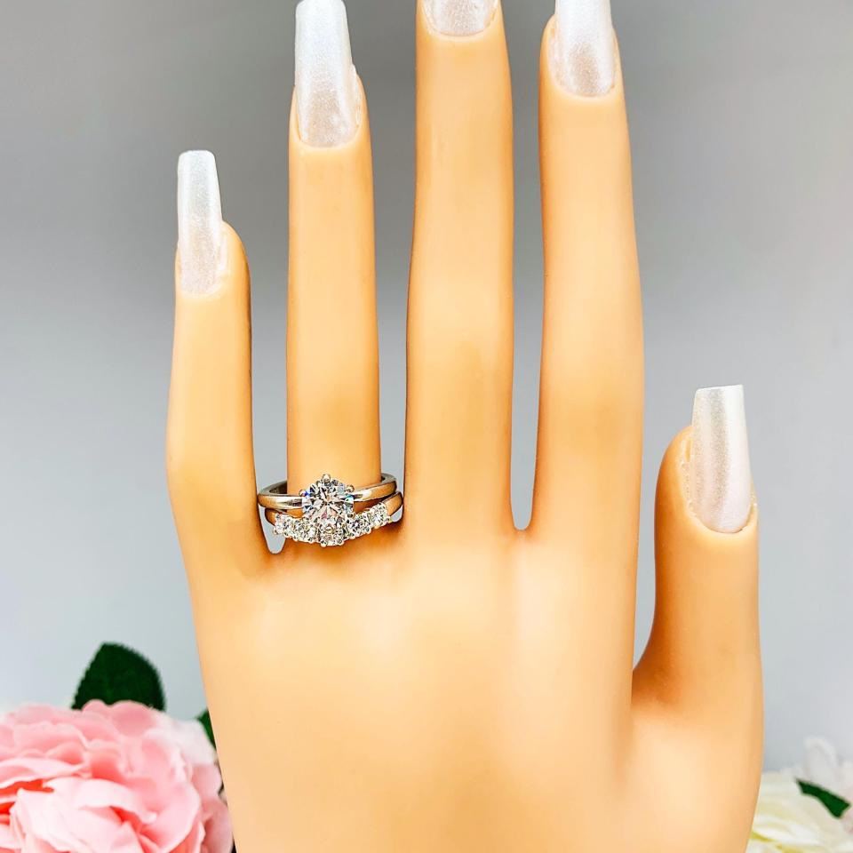 Mannequin hand with long white shimmery nails, displayed against a soft white-gray background with pink and white floral accents. The hand is wearing a sterling silver 5-stone curved wedding band featuring 3mm round precision faceted moissanite stones in prong settings. The ring is paired with a solitaire round cz engagement ring. This ring is not included.