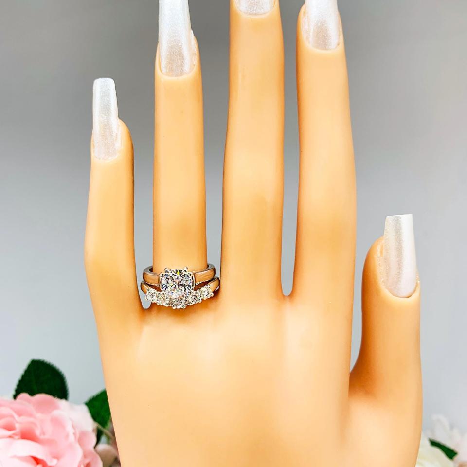 Mannequin hand with long white shimmery nails, displayed against a soft white-gray background with pink and white floral accents. The hand is wearing a sterling silver 5-stone curved wedding band featuring 3mm round precision faceted moissanite stones in prong settings. The ring is paired with a solitaire cushion cz engagement ring. This ring is not included.