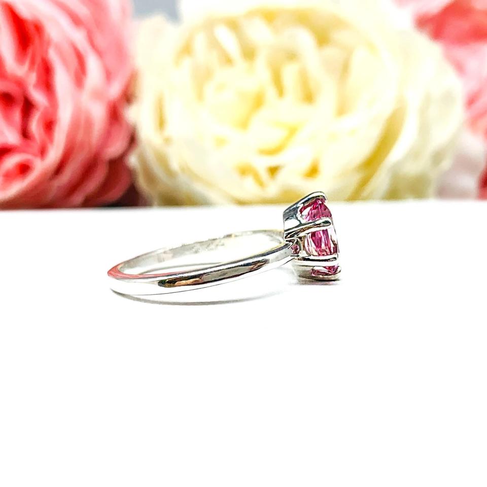 Sterling silver ring with an 8x6mm oval lab created padparadscha sapphire set in a 6-prong setting set against a white surface with soft-focus pink and white flowers in the background.