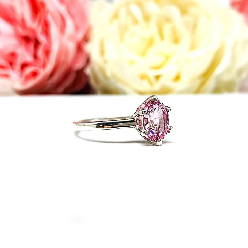 Sterling silver ring with an 8x6mm oval lab created padparadscha sapphire set in a 6-prong setting set against a white surface with soft-focus pink and white flowers in the background.