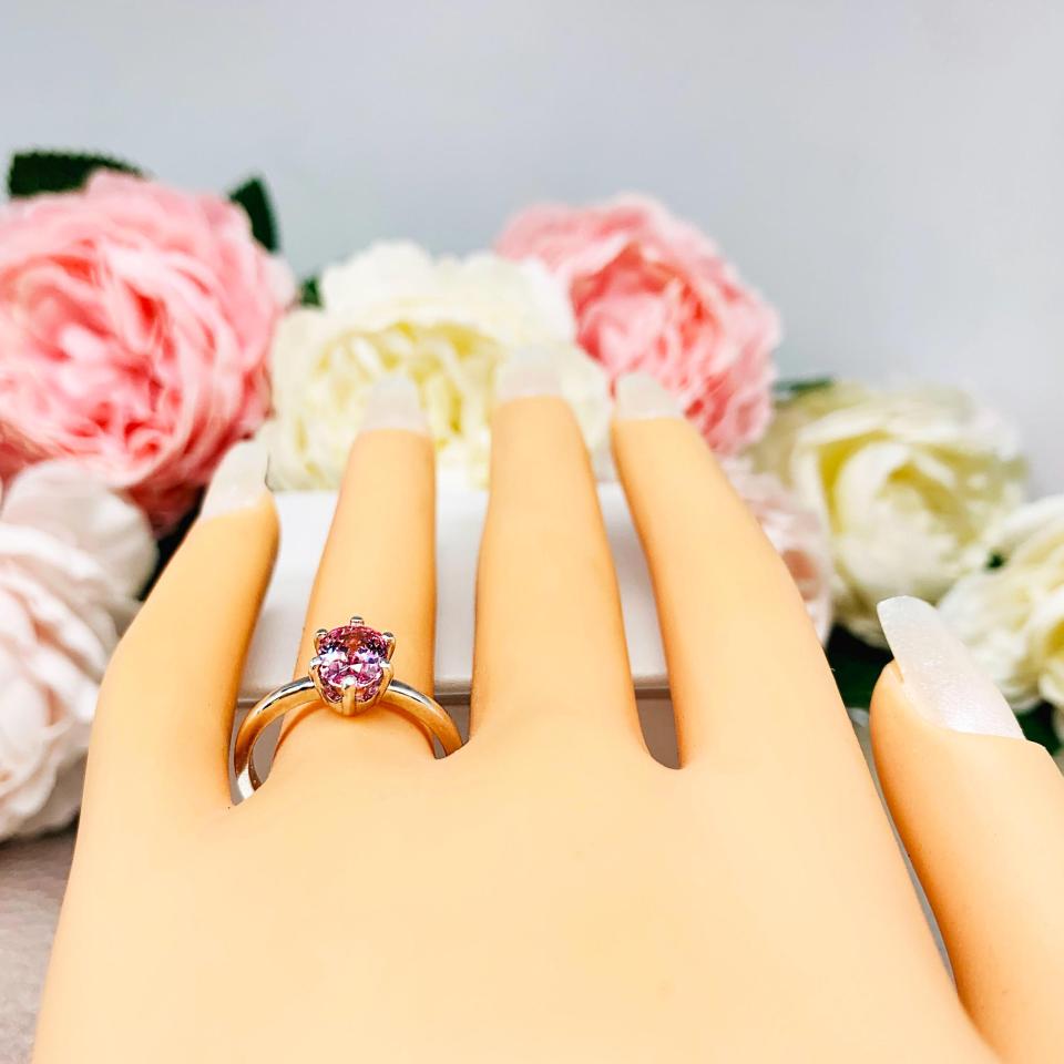 Mannequin hand with long white shimmery nails, displayed against a soft white-gray background with pink and white floral accents. The hand is wearing a sterling silver ring with an 8x6mm oval lab created padparadscha sapphire set in a 6-prong setting.