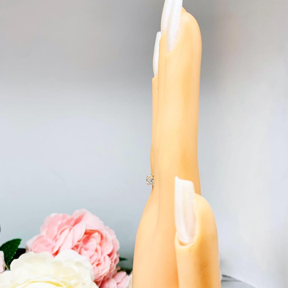 Mannequin hand with long white shimmery nails, displayed against a soft white-gray background with pink and white floral accents. The hand is wearing  a sterling silver graduated moissanite engagement ring featuring a 4mm center stone flanked by 3mm and 2.5mm side stones.