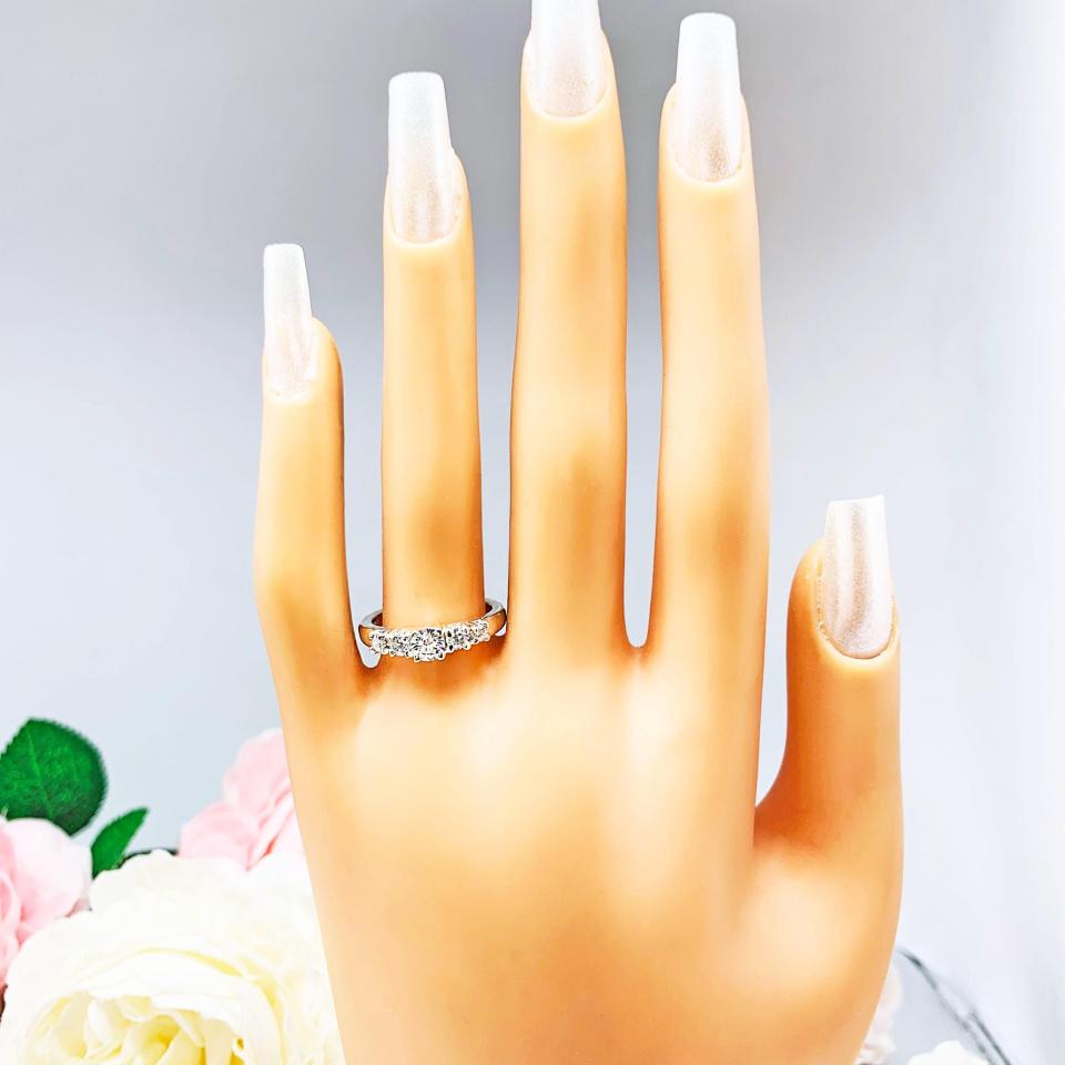 Mannequin hand with long white shimmery nails, displayed against a soft white-gray background with pink and white floral accents. The hand is wearing  a sterling silver graduated moissanite engagement ring featuring a 4mm center stone flanked by 3mm and 2.5mm side stones.