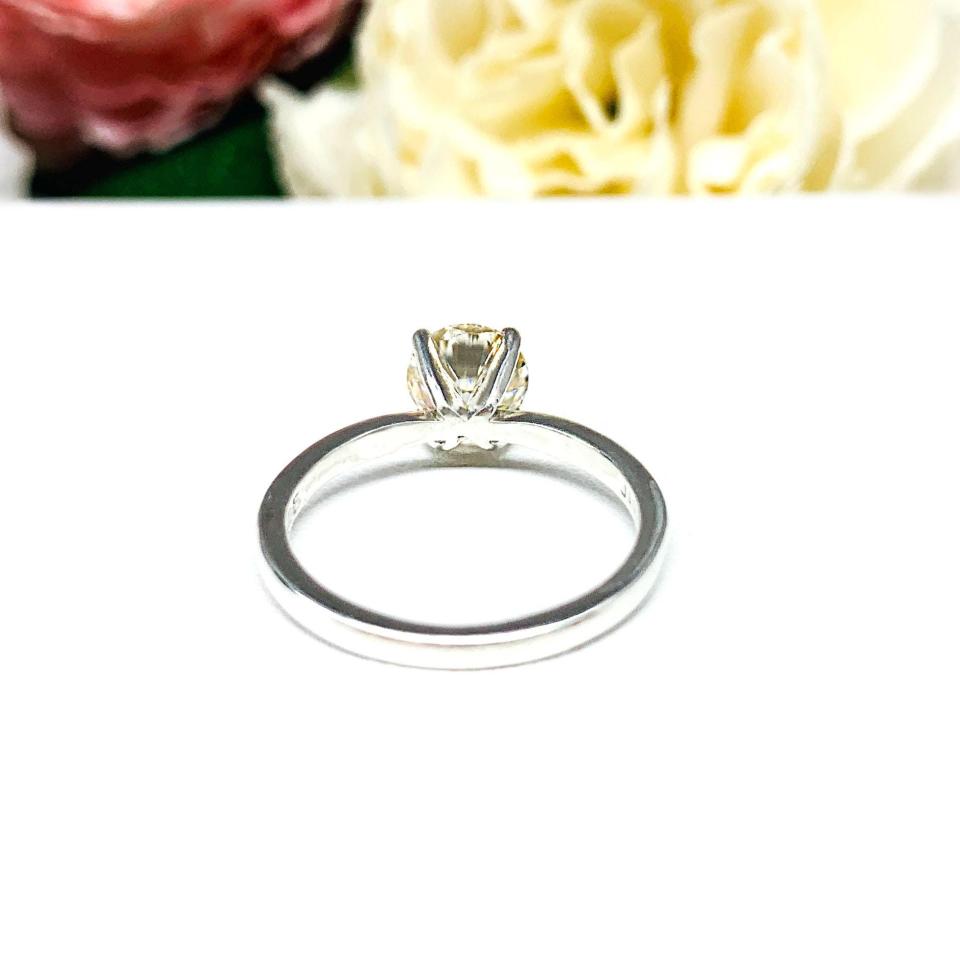 A solitaire engagement ring with a round brilliant light champagne moissanite. The setting is a classic four-prong design, and the band is a simple, sterling silver band. The ring is displayed on a white background with blurred pink and yellow flowers in the background.