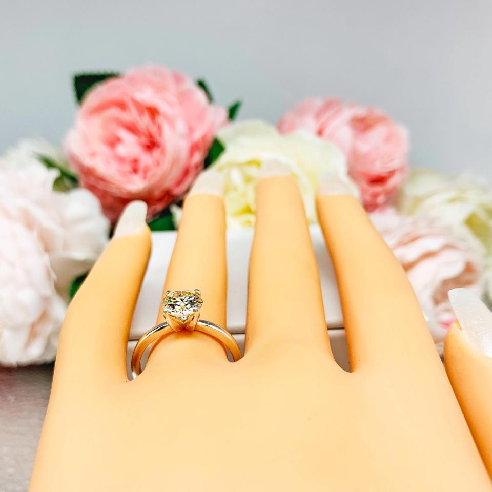 Mannequin hand with long white shimmery nails, displayed against a soft white-gray background with pink and white floral accents. The hand is wearing a solitaire engagement ring with a round brilliant light champagne moissanite. The setting is a classic four-prong design, and the band is a simple, sterling silver band.