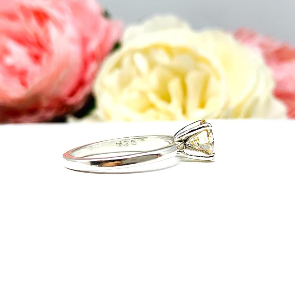 A solitaire engagement ring with a round brilliant light champagne moissanite. The setting is a classic four-prong design, and the band is a simple, sterling silver band. The ring is displayed on a white background with blurred pink and yellow flowers in the background.