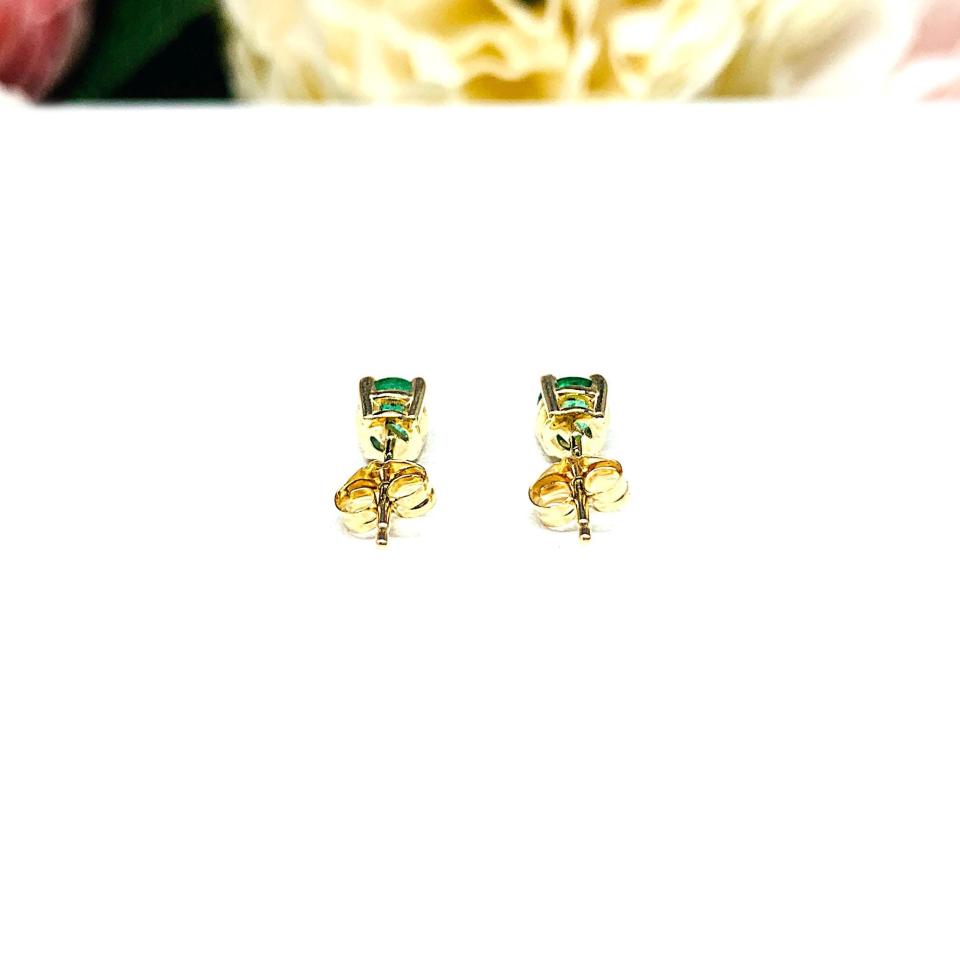 A pair of 14k yellow gold stud earrings set with 4mm round natural emerald gemstones. The four prong basket earrings are on a white surface with both earrings facing toward the back to showcase the center post and butterfly friction clasps.