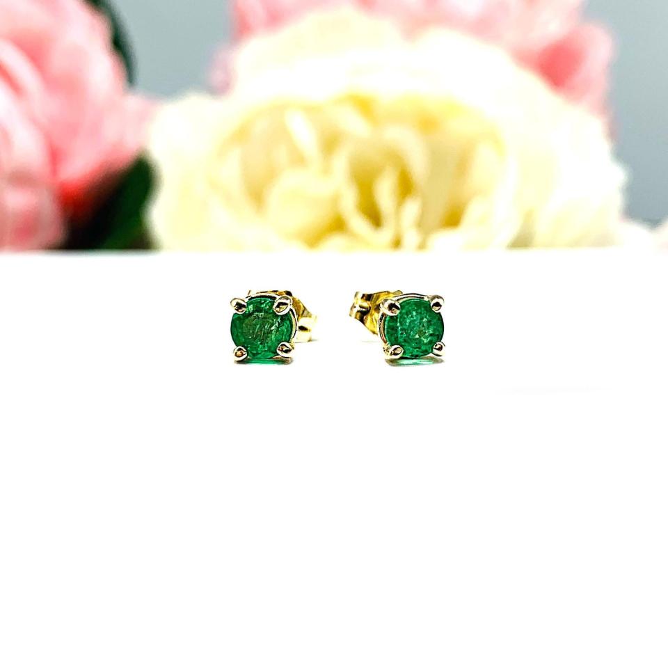 A pair of 14k yellow gold stud earrings set with 4mm round natural emerald gemstones. The four prong basket earrings are on a white surface with both earrings facing front to showcase the clarity and vibrant green color of the stones.