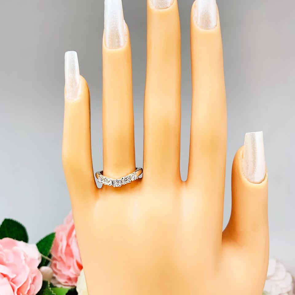 Mannequin hand with long white shimmery nails, displayed against a soft white-gray background with pink and white floral accents. The hand is wearing a sterling silver 5-stone curved wedding band featuring 3mm round precision faceted moissanite stones in prong settings.