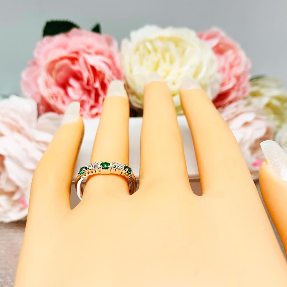 Mannequin hand with long white shimmery nails, displayed against a soft white-gray background with pink and white floral accents. The hand is wearing a sterling silver  five-stone curved wedding band with three 3mm round lab-created emeralds and two 3mm round DEF moissanites.
