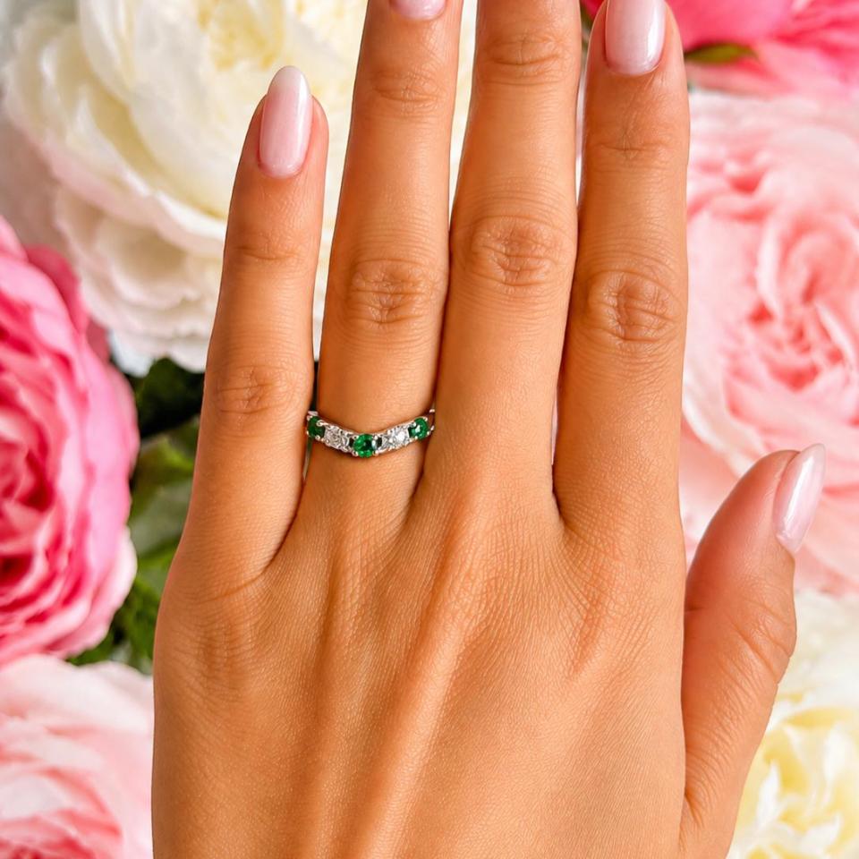 A woman's hand wearing a sterling silver five-stone curved wedding band with three 3mm round lab-created emeralds and two 3mm round DEF moissanites.