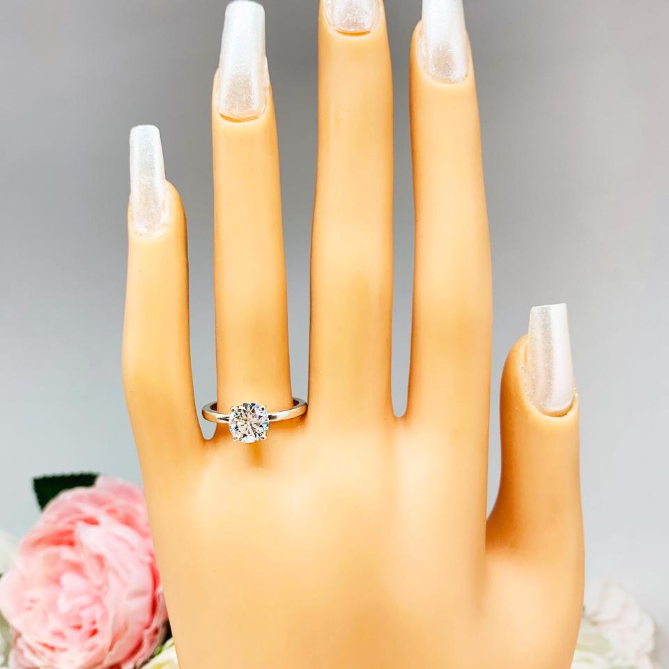 Mannequin hand with long white shimmery nails, displayed against a soft white-gray background with pink and white floral accents. The hand is wearing a solitaire engagement ring with a round brilliant DEF moissanite. The setting is a classic four-prong design, and the band is a simple, sterling silver band. The ring is displayed on a white background with blurred pink and yellow flowers in the background.