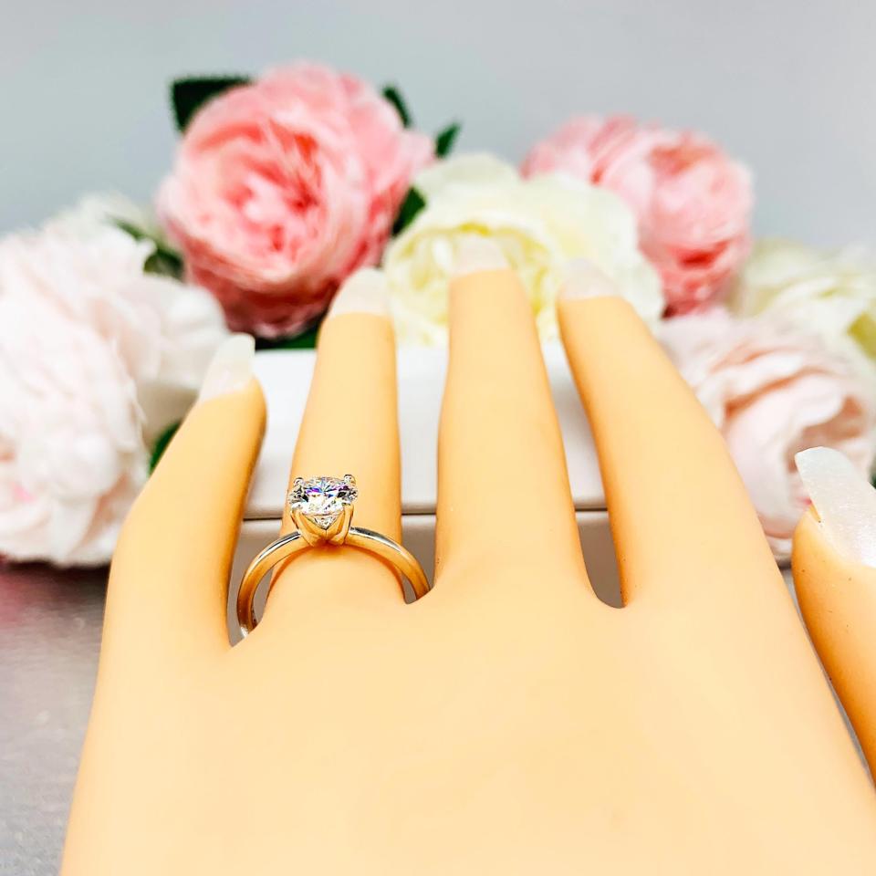 Mannequin hand with long white shimmery nails, displayed against a soft white-gray background with pink and white floral accents. The hand is wearing a solitaire engagement ring with a round brilliant DEF moissanite. The setting is a classic four-prong design, and the band is a simple, sterling silver band. The ring is displayed on a white background with blurred pink and yellow flowers in the background.