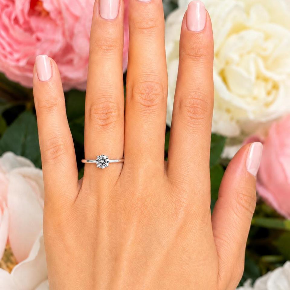 Woman's hand wearing a sterling silver 5mm round-cut 4-prong moissanite engagement ring. Model image for size and styling reference — not a photo of the actual ring.