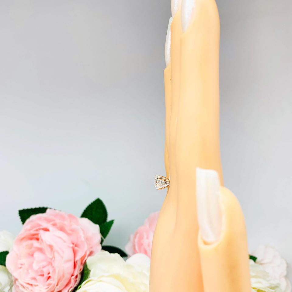 Mannequin hand with long white shimmery nails, displayed against a soft white-gray background with pink and white floral accents. The hand is wearing a sterling silver 4-prong asscher cut DEF moissanite solitaire engagement ring.