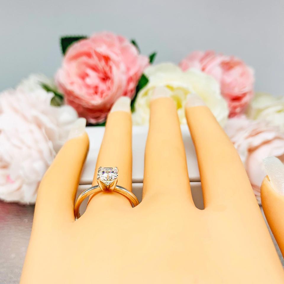 Mannequin hand with long white shimmery nails, displayed against a soft white-gray background with pink and white floral accents. The hand is wearing a sterling silver 4-prong asscher cut DEF moissanite solitaire engagement ring.