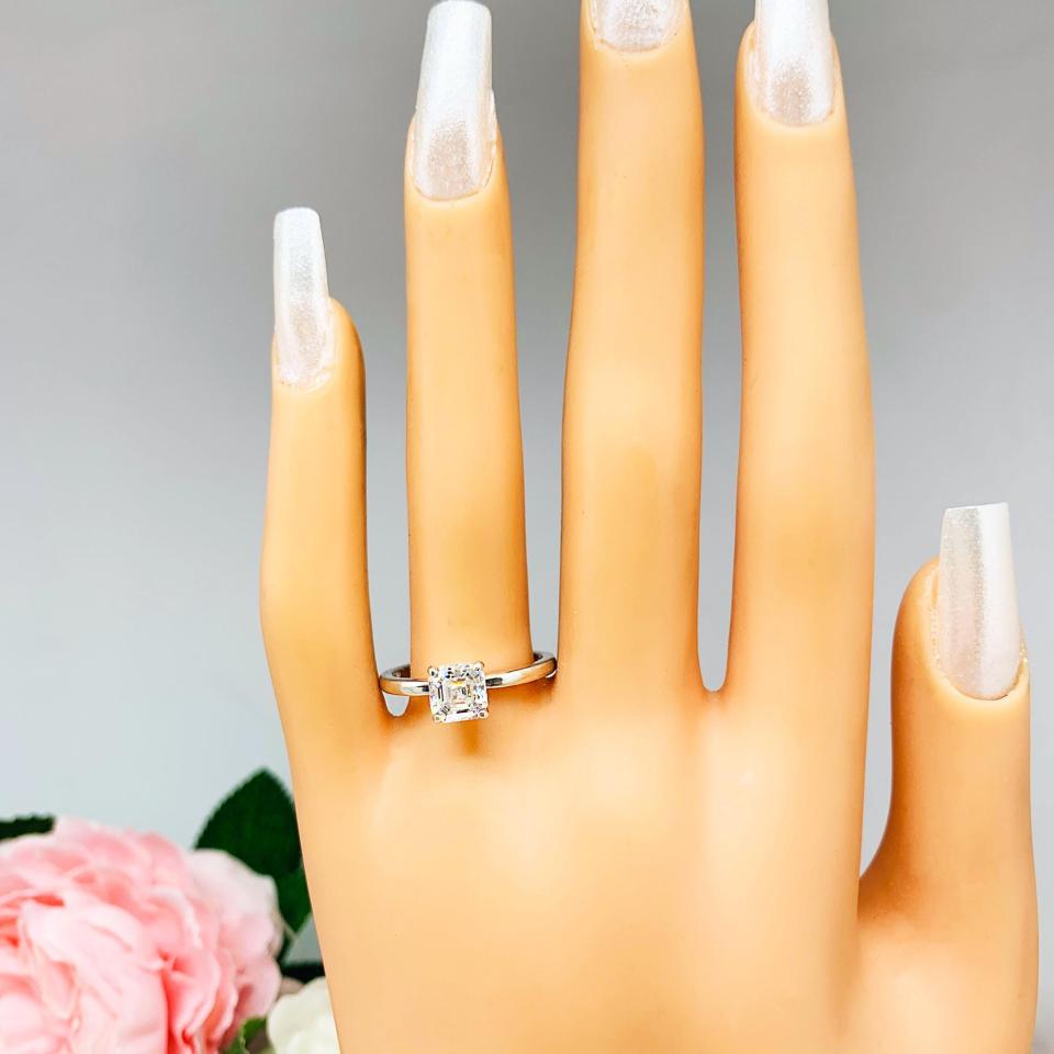 Mannequin hand with long white shimmery nails, displayed against a soft white-gray background with pink and white floral accents. The hand is wearing a sterling silver 4-prong asscher cut DEF moissanite solitaire engagement ring.