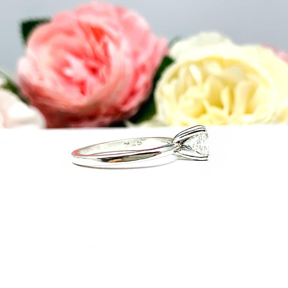 Sterling silver 4-prong asscher cut DEF moissanite solitaire engagement ring. The ring is resting on a white surface with a background of pink and white flowers.